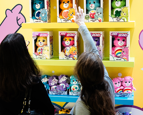 Two individuals browsing a colorful display of Care Bears plush toys in boxed packaging at Brand Licensing Europe